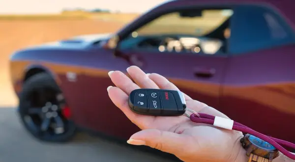 Kluczyk do nowego auta w dłoni. To pierwszy krok w kupnie samochodu krok po kroku. Czerwony Dodge Challenger w tle.