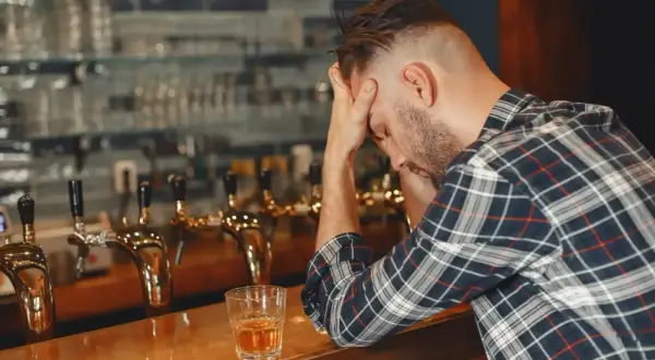 Mężczyzna z rękami na głowie siedzi przy barze, obok szklanka z alkoholem. Może to być jeden z 6 objawów uzależnienia od alkoholu.