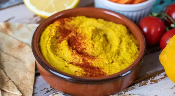 En skål med krämig hummus, toppad med paprikapulver, serveras med pitabröd, citron och grönsaker. Perfekt för ett enkelt hummus recept.