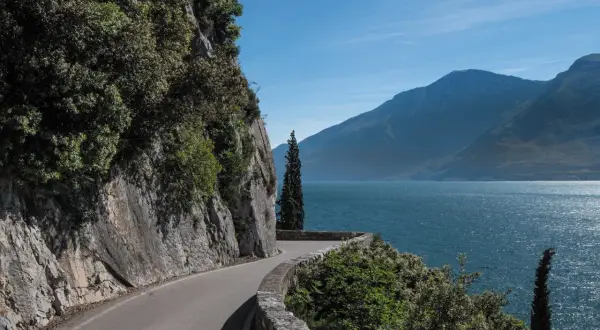 Strada panoramica lungo il lago, con rocce a sinistra e montagne sullo sfondo. La gardesana occidentale offre scorci mozzafiato.