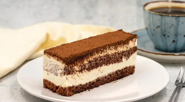Pyszne ciasto czekoladowe z mascarpone, posypane kakao, na białym talerzu, obok filiżanka kawy.