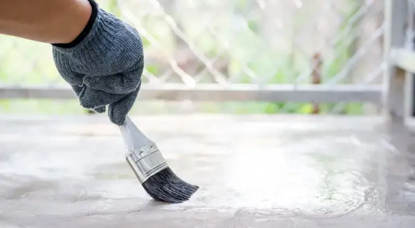 Ręka w rękawicy nakłada pędzelkiem bezbarwny preparat na betonową posadzkę. To dobry sposób, czym pomalować beton, by go zabezpieczyć.