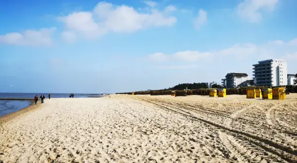 Ein weiter, sonniger Duhnen Strand mit gelben Strandkörben und einigen Spaziergängern. Im Hintergrund moderne Gebäude.