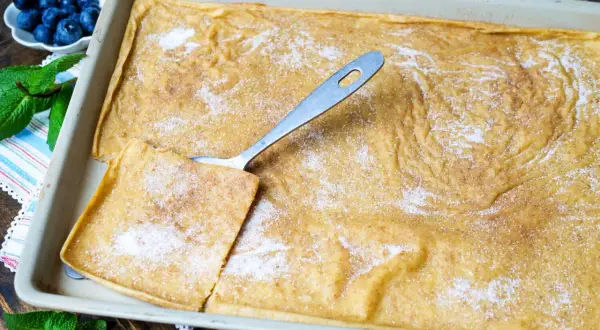Großer, goldbrauner Pfannkuchen vom Blech, bestreut mit Zucker und Zimt. Ein Stück wird mit einem Löffel herausgehoben. Blaubeeren und Minze garnieren.