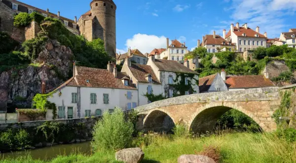 Un village pittoresque de la Bourgogne avec un pont de pierre ancien, des maisons aux toits rouges et une tour médiévale perchée sur une colline verdoyante.