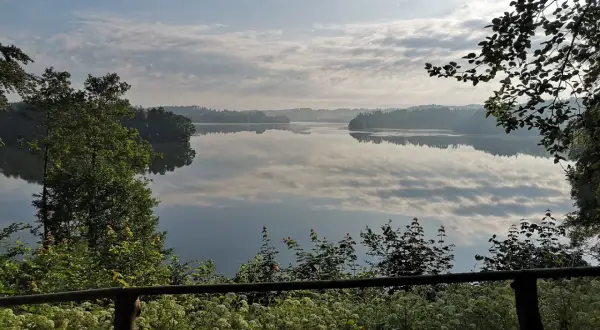 Spokojny poranek nad jeziorem w Sulęczynie. Lustrzana tafla wody odbija chmury i zalesione brzegi.