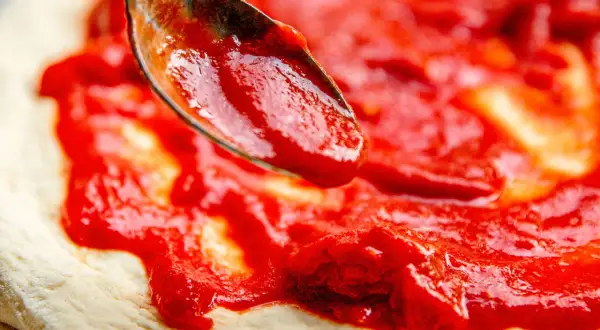 Ein Löffel verteilt die leuchtend rote San Marzano Pizza Sauce auf einem frischen Pizzateig.