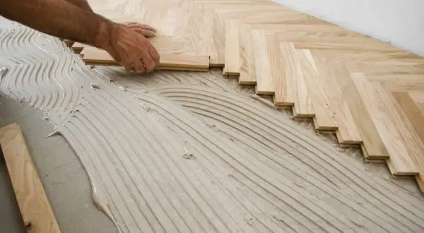 Mains d'un artisan en train de poser du parquet en chevrons sur un lit de colle ondulée.