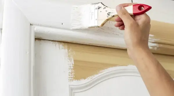 Une main applique de la peinture blanche sur une porte intérieure en bois. Le travail de peinture est en cours, révélant le bois naturel sous la couche blanche.