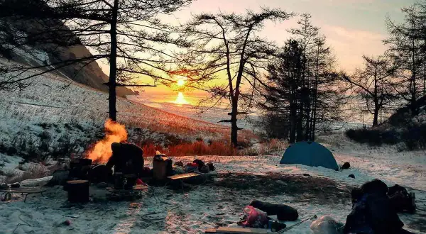 Campamento invernal con fogata y tienda de campaña. La puesta de sol ilumina el paisaje nevado, un ejemplo de supervivencia y naturaleza.