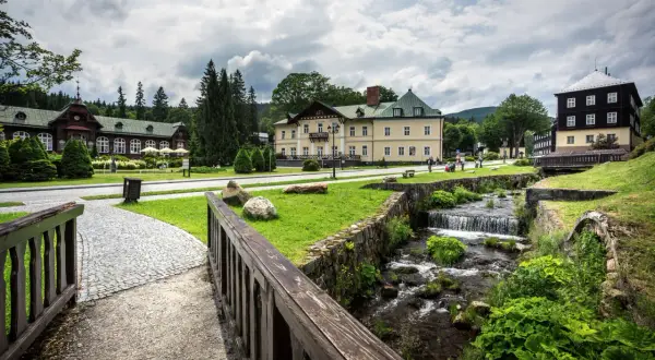 Idyllische Szene in Karlova Studanka: Gebäude im Kurort, ein Bach plätschert über Steine, umgeben von Grün.