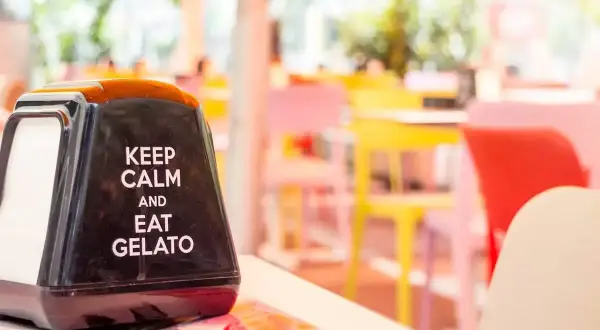 Na stoliku stoi czarny dozownik na serwetki z napisem "Keep calm and eat gelato". W tle kolorowe krzesła w kawiarni. To dobre hasło na lato!