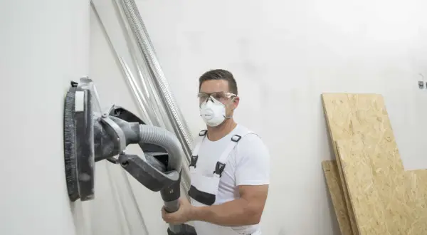Operaio con maschera e occhiali sta per carteggiare muro senza fare polvere, usando un aspiratore integrato.