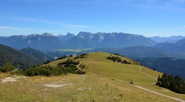 Ein sonniger Tag lädt zum **Wank wandern** ein. Grüne Hügel mit verschlungenen Pfaden erstrecken sich vor einer majestätischen Bergkulisse.