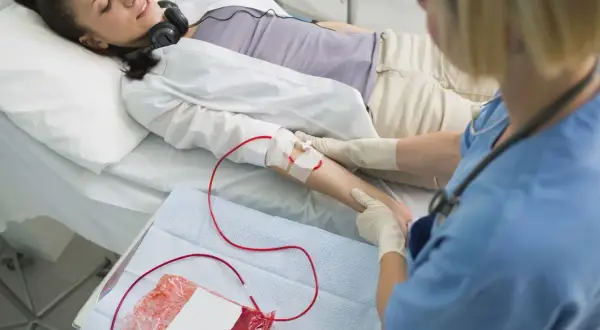 Kobieta leży w łóżku, słuchając muzyki przez słuchawki, podczas gdy pielęgniarka wykonuje przetaczanie krwi.