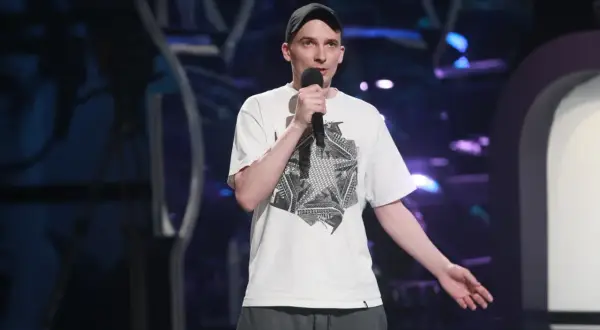 Jasiek Borkowski w czapce i białej koszulce z grafiką, trzymający mikrofon, podczas występu stand-up.