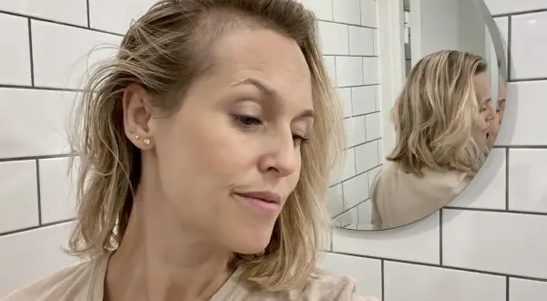 Kvinna med blont hår tittar ner, reflekteras i spegeln. Hon verkar orolig över att tappa hår under amning.