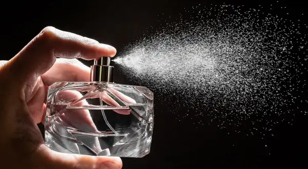 Dłoń trzyma szklaną buteleczkę, z której rozpyla się mgiełka. To woda perfumowana, a nie perfumy, tworząca subtelny zapach.