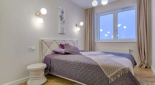 Une chambre moderne aux murs clairs, prête à peindre. Un lit douillet avec des coussins violets et une couverture à motifs. Des appliques murales design et une fenêtre lumineuse.