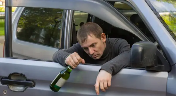 Mężczyzna z butelką w ręku opiera się o otwarte drzwi samochodu. Jego mina sugeruje, że może być to jazda pod wpływem.