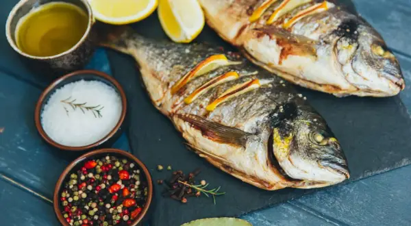 Zwei gebratene Dorade Fische mit Zitronenscheiben, Olivenöl, Salz und Pfefferkörnern auf einem dunklen Brett.