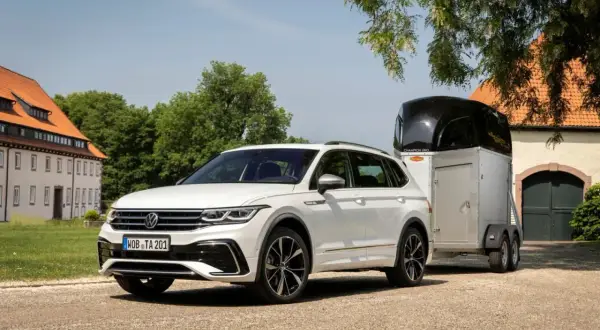 Biały Volkswagen Tiguan, jeden z wielu wszechstronnych volkswagen modele, holuje przyczepę z koniem przed zabytkowym budynkiem.