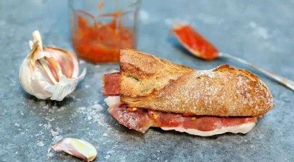A delicious bocadillo recipe featuring a crusty baguette filled with cured ham and a vibrant tomato spread, with garlic and a jar of spread nearby.