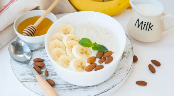 Pyszna owsianka proteinowa z bananem, migdałami i miętą. Obok miseczka z miodem i dzbanuszek z mlekiem.