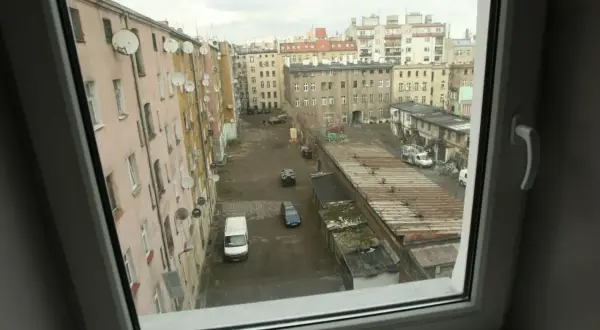 Podwórko kamienicy z satelitami na ścianach. Widać samochody i garaże. Trudno ocenić, ile metrów na osobę mieszkanie komunalne.