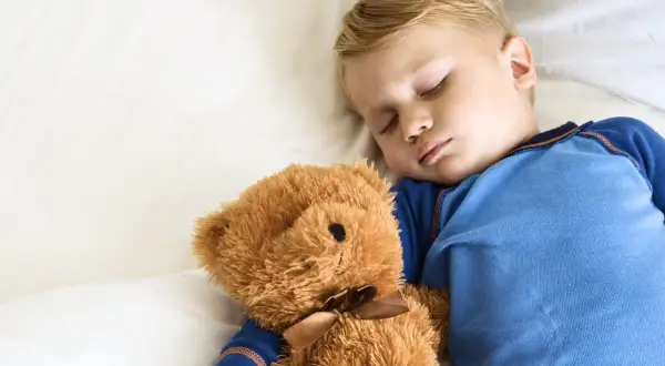 En liten pojke sover sött med sin nallebjörn. Hur många timmar ska en 10-åring sova? Sömn är viktigt för barn.