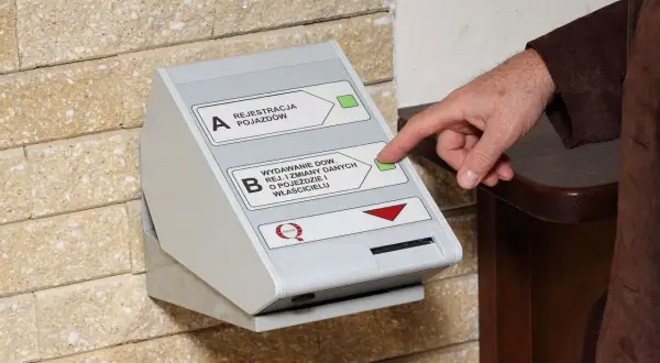 Dłoń naciska przycisk na automacie do wydawania dowodów rejestracyjnych, ułatwiając wymianę dowodu rejestracyjnego.