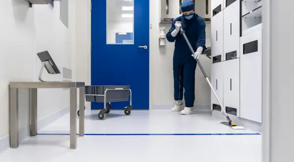 Osoba w kombinezonie i masce myje podłogę w sterylnym, clean room. W tle niebieskie drzwi i białe szafki.