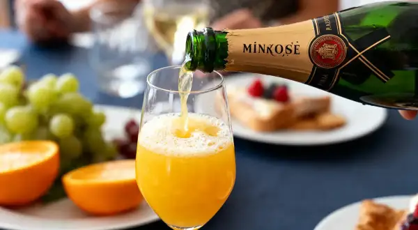 Une bouteille de Mimosé verse son contenu pétillant dans un verre rempli de jus d'orange. Un brunch festif se prépare.