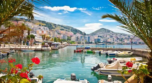 Albania, gdzie najpiękniej! Widok na port z palmami, kolorowymi budynkami na wzgórzu i łodziami na turkusowej wodzie.