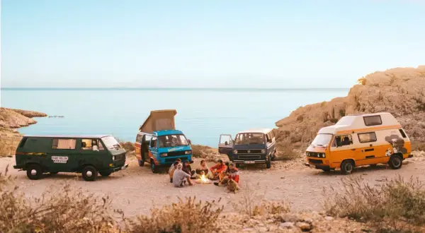 Vier bunte Campervans am Meer, Menschen sitzen am Lagerfeuer.