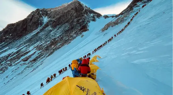 Eine lange Schlange von Bergsteigern kämpft sich den schneebedeckten Hang des Mount Everest hinauf. Die Frage, wie lange dauert es den Mount Everest zu besteigen, wird hier durch die schiere Anzahl der Menschen beantwortet.