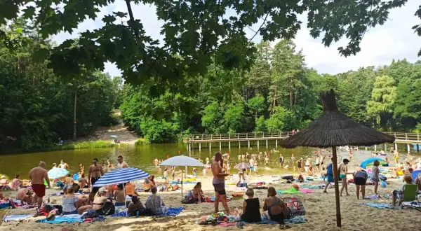 Słoneczny dzień na plaży przy zalewie w Garbatce. Ludzie odpoczywają na piasku, kąpią się w wodzie i spacerują po pomoście.