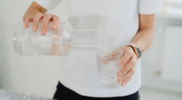 En person häller upp vatten från en glasflaska till ett glas. En hälsosam dryck som mot rethosta.