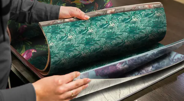 Händer bläddrar i en tapetbok med djungelmotiv och blomstermönster. Perfekt för att välja non woven lim till papperstapet.