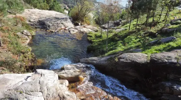 Cascada y pozas naturales en Pozas da Moura, con agua cristalina cayendo sobre rocas y vegetación exuberante. Al fondo, un estuario y colinas verdes.