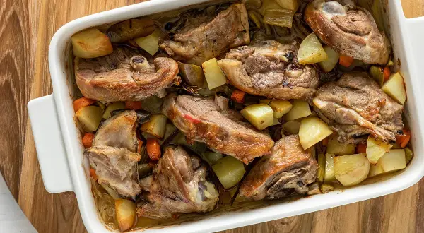 Capretto al forno ricetta della nonna: succulenti pezzi di carne dorata con patate e carote in una teglia bianca.
