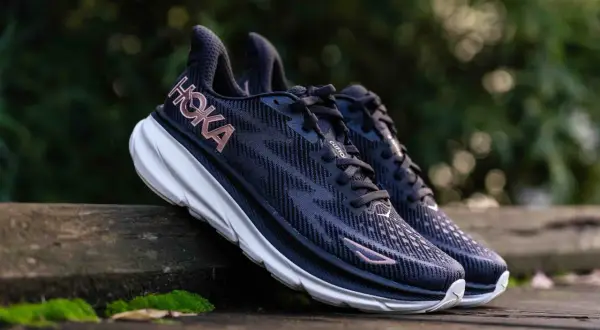 Paire de chaussures de course Hoka Clifton 9, bleues foncées avec logo rose, posées sur un banc en bois. Avis sur le confort et la performance.