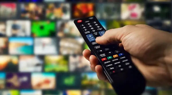 Dłoń trzymająca pilot do telewizora na tle wielu ekranów.