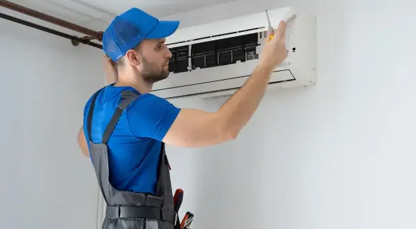 Technik w niebieskiej czapce i szarym ogrodniczkach naprawia klimatyzator śrubokrętem.