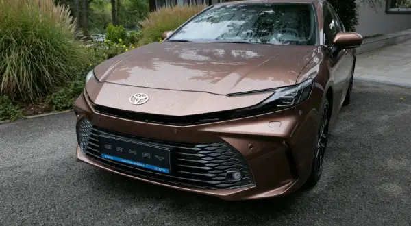 Nowa Toyota Camry 2024 w kolorze brązowym, z kroplami deszczu na karoserii, zaparkowana przed nowoczesnym budynkiem.