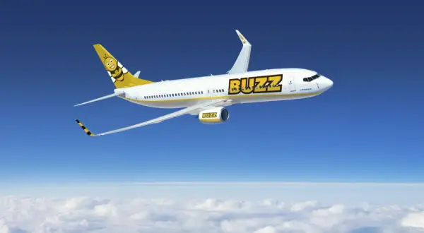 Samolot linii Buzz, należącej do Ryanair, z uśmiechniętą pszczołą na ogonie, leci na tle błękitnego nieba i chmur.