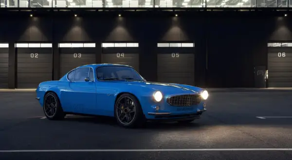 Niebieskie Volvo P1800 z nowoczesnymi felgami stoi przed budynkiem z numerowanymi bramami.