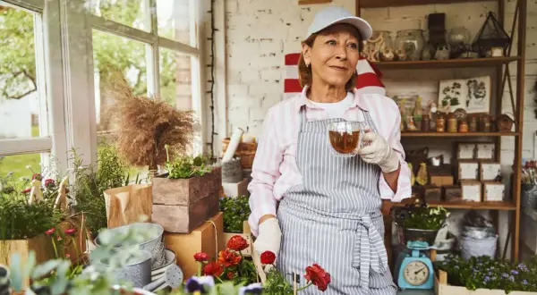 Frau mit Kappe und Schürze trinkt Tee im Blumenladen. Sie genießt ihre Zeit, vielleicht im Rahmen von altersteilzeit modellen, umgeben von Pflanzen.
