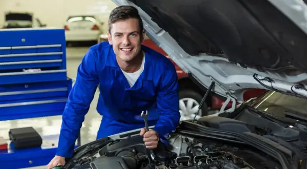 Uśmiechnięty mechanik w niebieskim kombinezonie z kluczem w dłoni, pracujący przy silniku. Dowiedz się, jak zostać elektrykiem samochodowym.