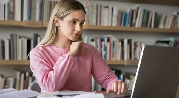 Dziewczyna w różowej bluzie uczy się przy laptopie na tle regałów z książkami.